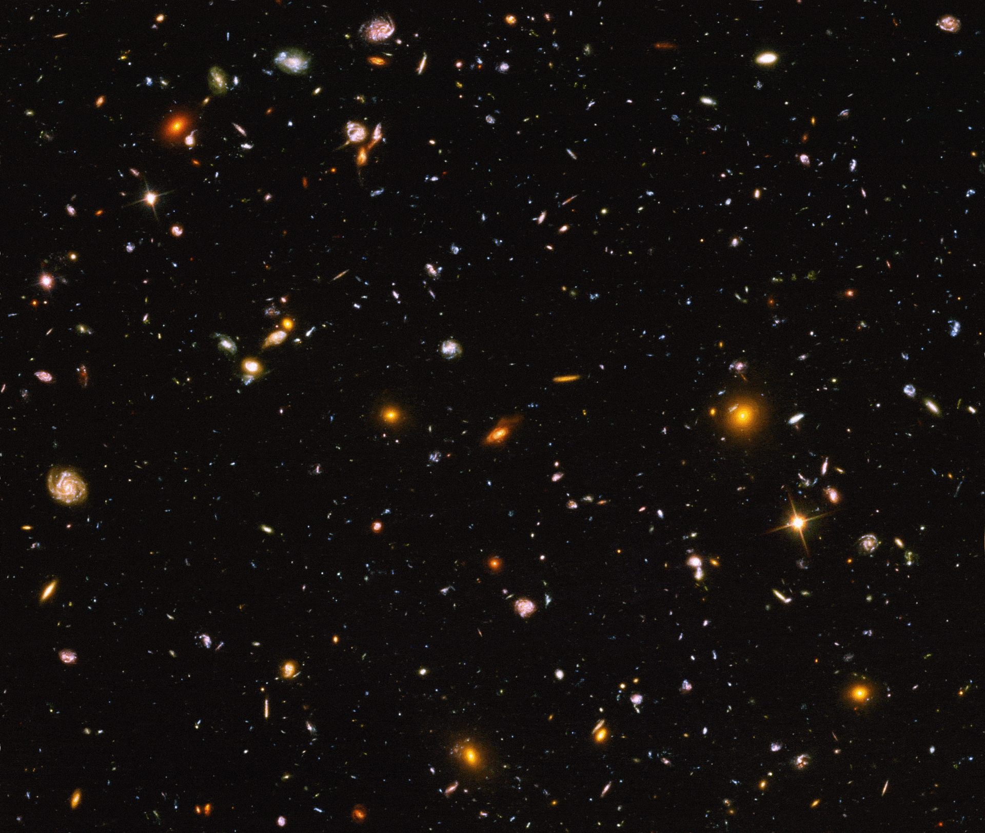 An area of deep space with thousands of galaxies in various shapes and sizes on a black background. Most are circles or ovals, with a few spirals. More distant galaxies are smaller, down to being mere dots, while closer galaxies are larger and some appear to be glowing. Red and orange galaxies contain more dust or more stellar activity. In the JWST verison everything is a lot brighter and a lot more galaxies are clearly visible.